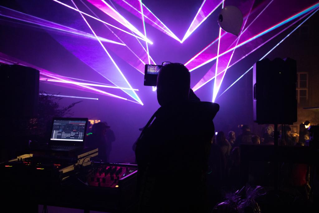 Blick über die Schulter des DJs am Pult mit Laptop, dahinter lila Laserstrahlen