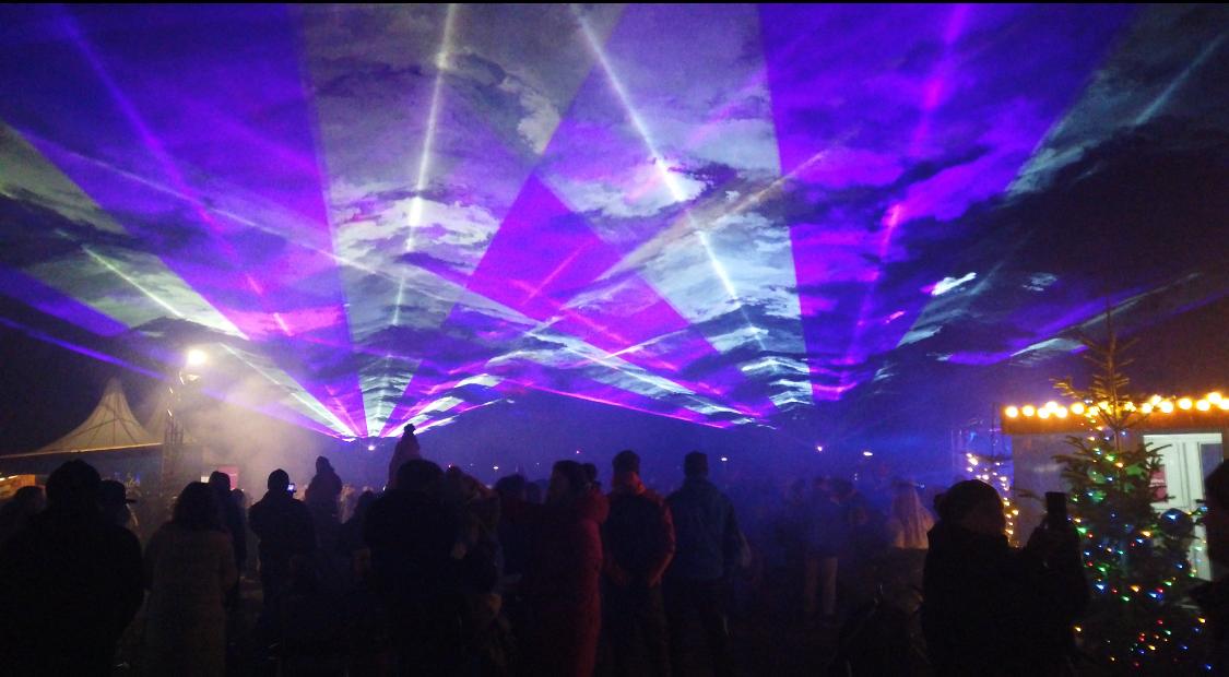 Lila und weiße Laserstrahlen mit Wolkenmuster über dem Publikum auf einem weihnachtlich beleuchteten Platz